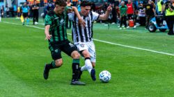 Adegan aksi dinamis dua pemain sepak bola yang berlomba untuk menguasai bola di lapangan.