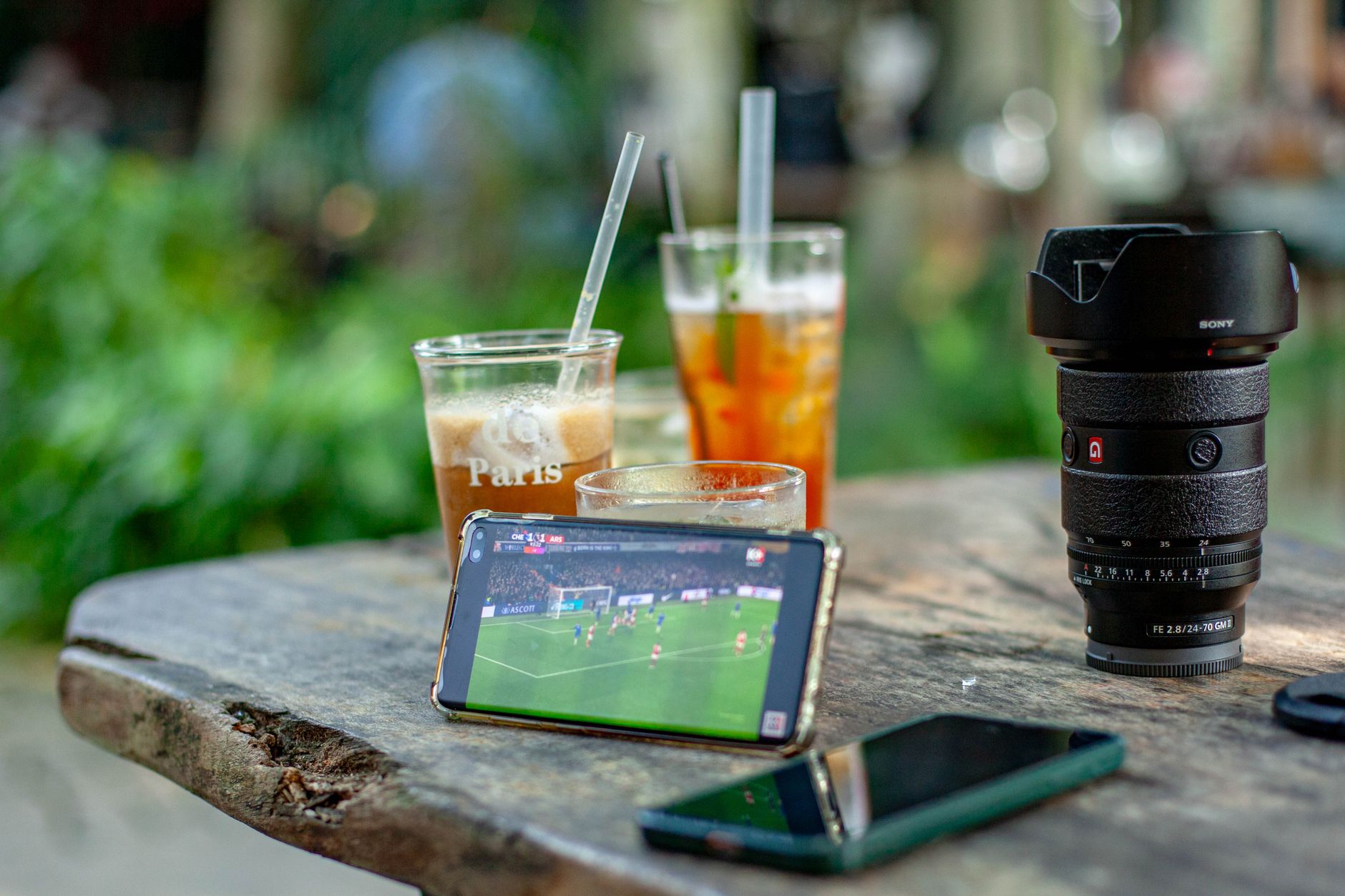 Sebuah telepon pintar menayangkan pertandingan sepak bola di samping minuman dan perlengkapan kamera di sebuah kafe luar ruangan.