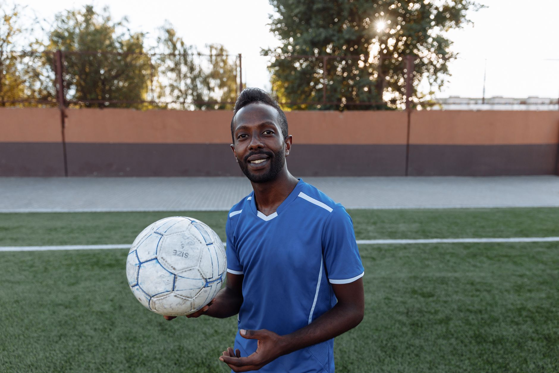 Seorang pemain sepak bola yang ceria mengenakan jersey biru memegang bola sepak di lapangan rumput, siap untuk bermain.