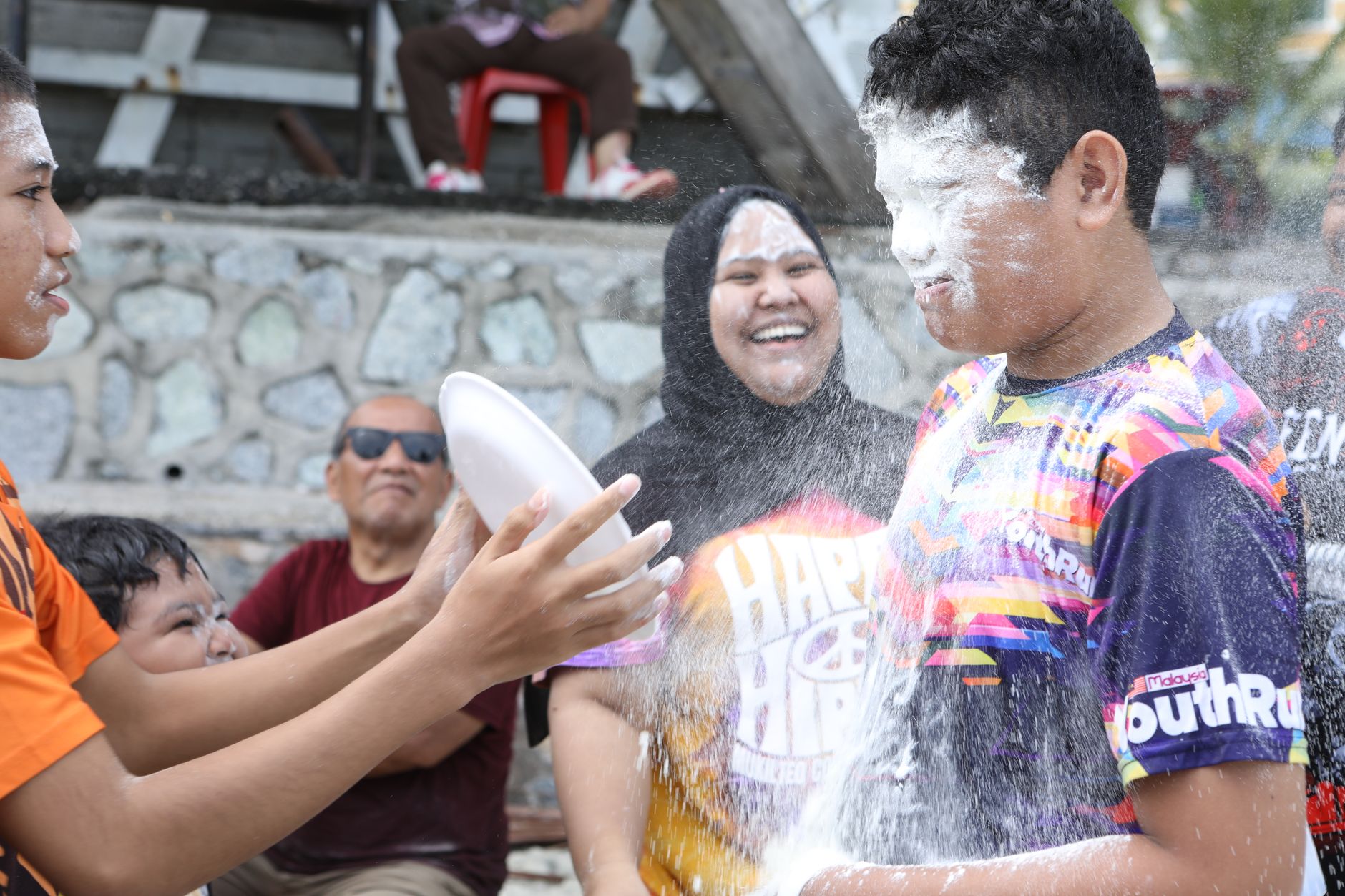 Sekelompok orang menikmati perayaan yang penuh permainan dengan tepung di luar ruangan, menunjukkan kebahagiaan dan kesenangan di Port Dickson, Malaysia.