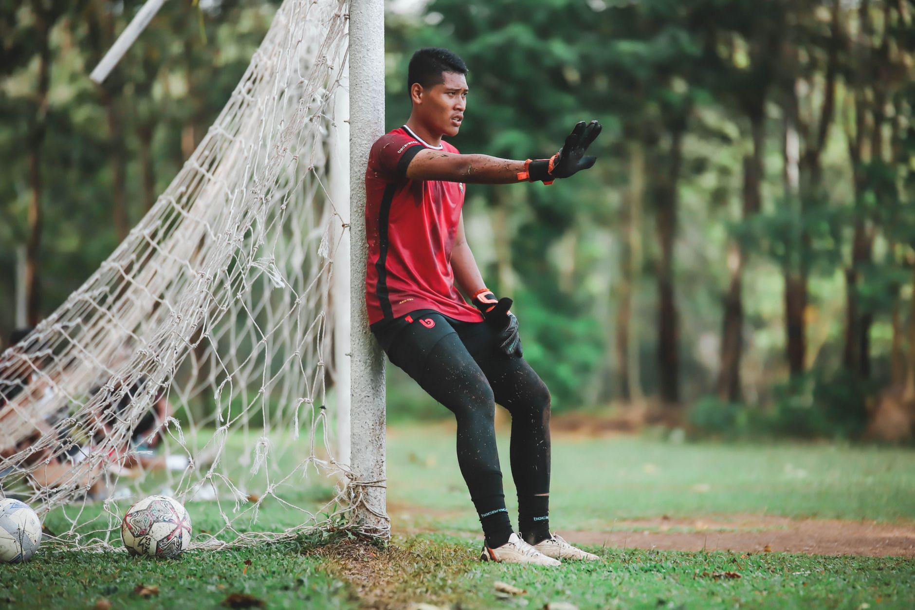 Seorang pemain sepak bola beristirahat di dekat tiang gawang di sebuah taman, mengenakan jersey merah.