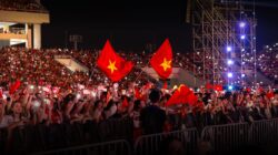 Sebuah konser malam dengan kerumunan besar yang energik melambaikan bendera di bawah lampu yang terang.
