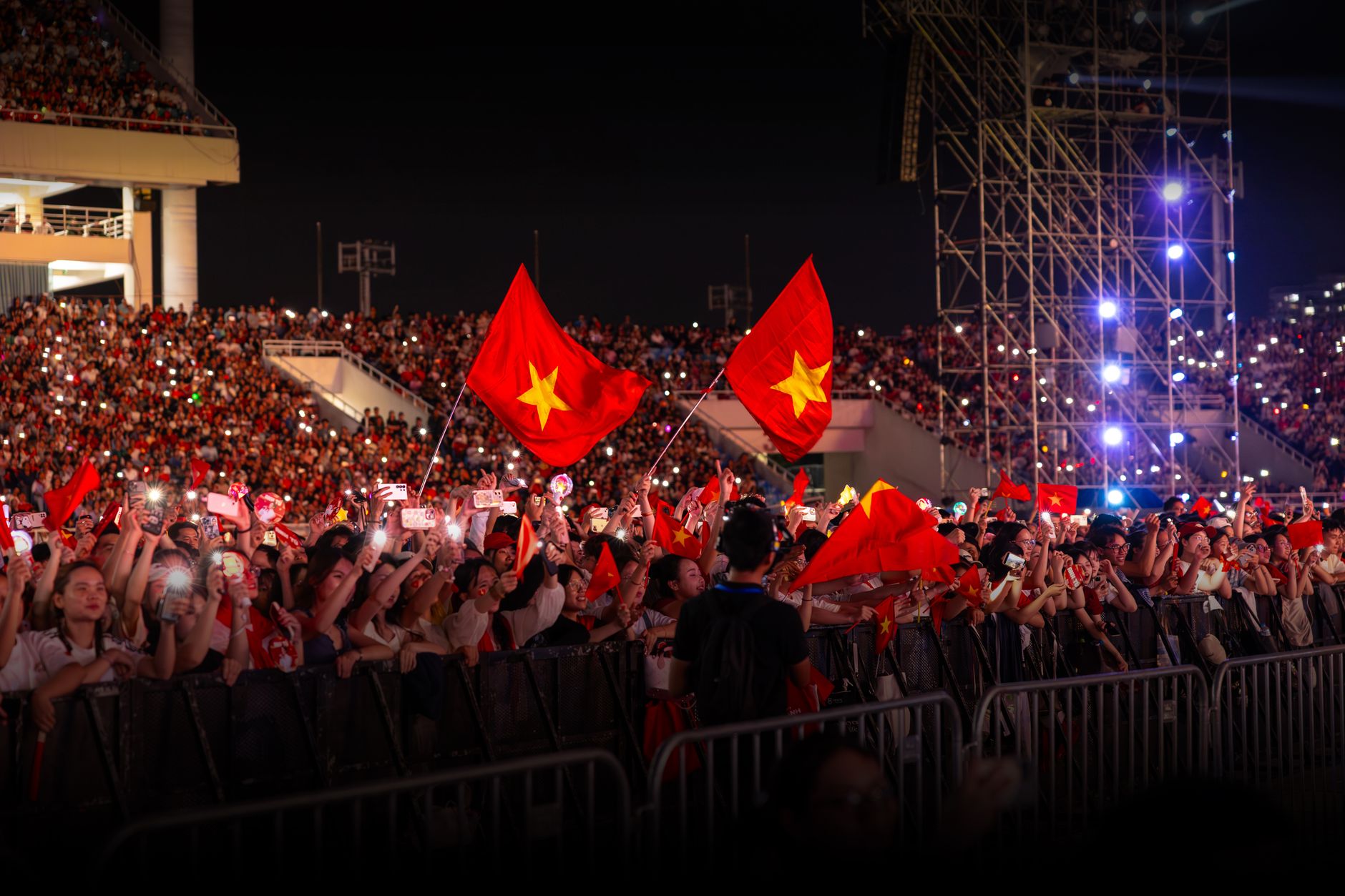 Sebuah konser malam dengan kerumunan besar yang energik melambaikan bendera di bawah lampu yang terang.
