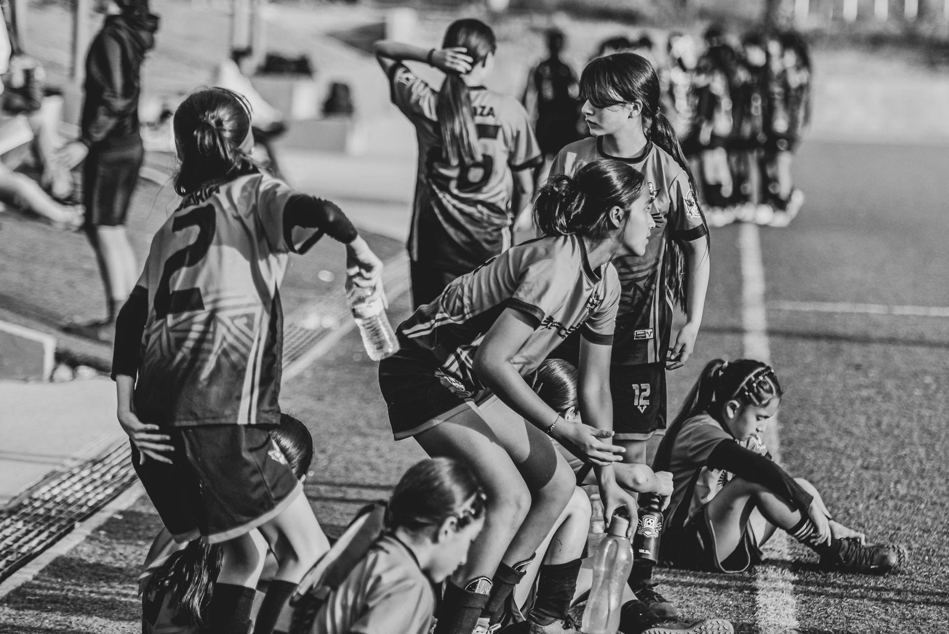 Tim sepak bola remaja perempuan beristirahat di sisi lapangan selama pertandingan dalam hitam dan putih.