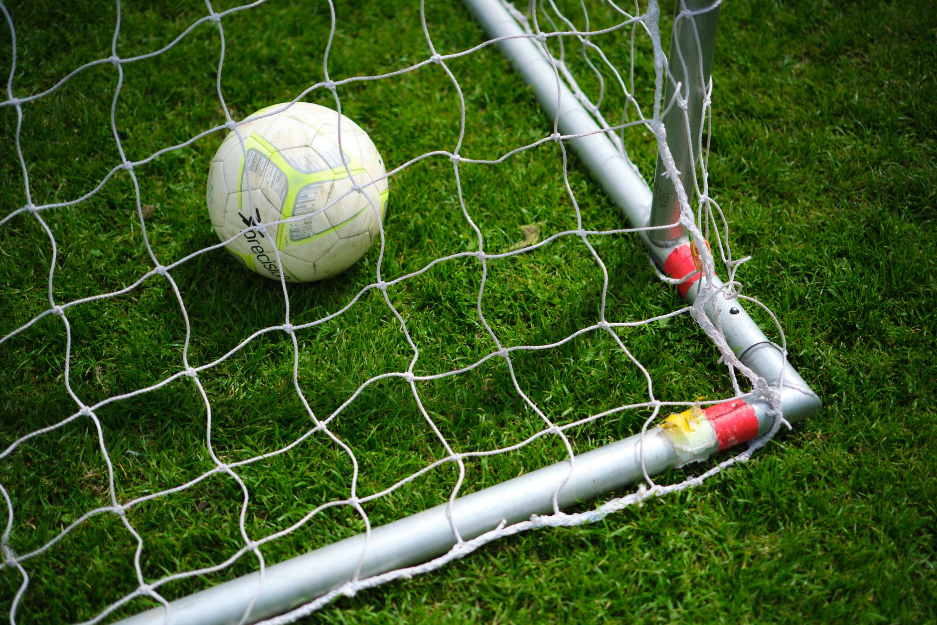 Gambar close-up sebuah bola sepak yang berada di dalam jaring gawang di lapangan rumput hijau yang cerah.