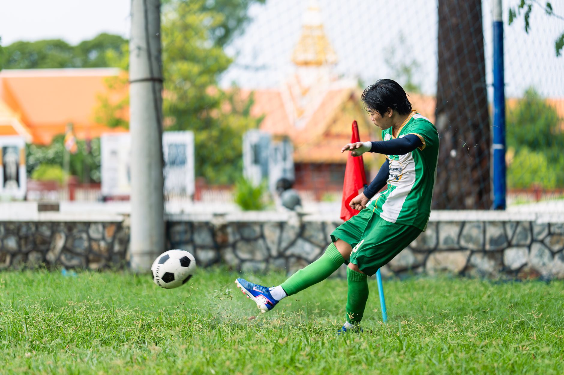 Gambar dinamis seorang pemain sepak bola muda menendang bola di lapangan terbuka pada siang hari.