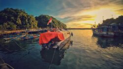Pemandangan matahari terbenam yang menakjubkan di Pelabuhan Girisubo, Indonesia, dengan perahu dan bukit hijau yang subur.