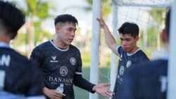 Dua pemain sepak bola mendiskusikan strategi di lapangan dekat tiang gawang di luar ruangan.