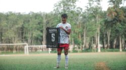 Seorang pemain sepak bola muda dengan bangga memegang jersey tim di lapangan hijau yang dikelilingi pohon.