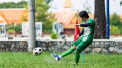 Gambar dinamis seorang pemain sepak bola muda menendang bola di lapangan terbuka pada siang hari.
