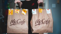 Seseorang memegang dua kantong pengantaran makanan McCafé di dalam ruangan, menekankan layanan makanan cepat saji.